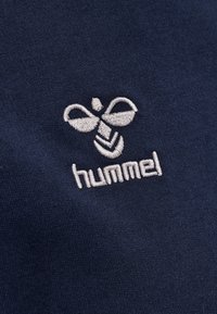 Tessuto blu navy con un logo ricamato di un'ape stilizzata sopra la parola "hummel" in filo grigio chiaro. Texture liscia.