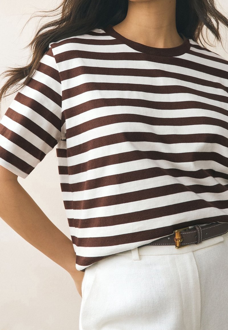 Femme portant un t-shirt à manches courtes marron et blanc à rayures horizontales, rentré dans un pantalon blanc avec une ceinture foncée.