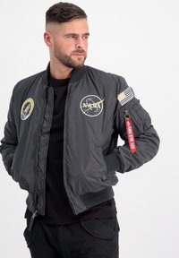 Giacca bomber grigia con logo NASA, toppe e etichetta rossa. Polsini e orlo a coste, chiusura con zip, con un aspetto lucido e slanciato.