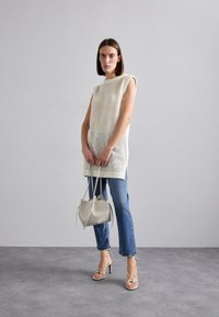 Tunika senza maniche in beige chiaro, abbinata a jeans blu e sandali con tacco a listini. Accompagnata da una piccola borsa a tracolla grigia chiara.