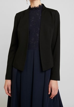 Zwarte getailleerde blazer met een gladde textuur, open voorkant en lange mouwen, gecombineerd met een marineblauwe jurk met een kantenkraag en vloeiende stof.