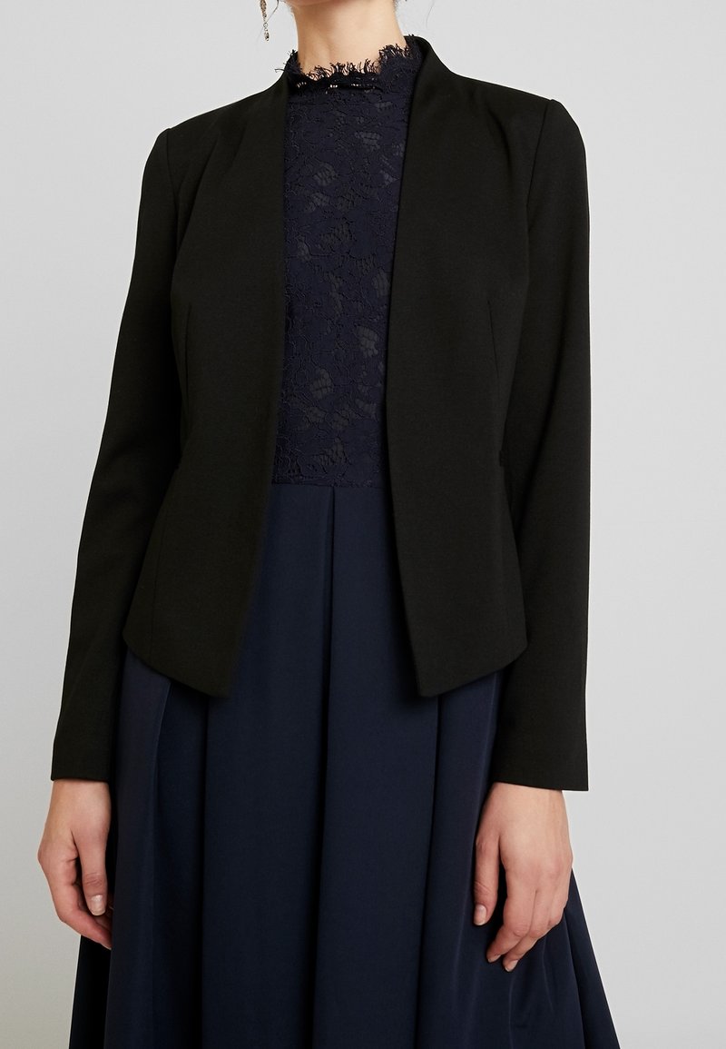 Musta istuva bleiseri, jossa on sileä tekstuuri, avonainen etuosa ja pitkät hihat, yhdistettynä tummansiniiseen mekkoon, jossa on pitsikaulus ja virtaava kangas.