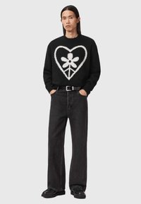 Svart fluffy genser med et stort hvitt hjerte og blomstermønster, kombinert med løstsittende mørke jeans og svarte sko.