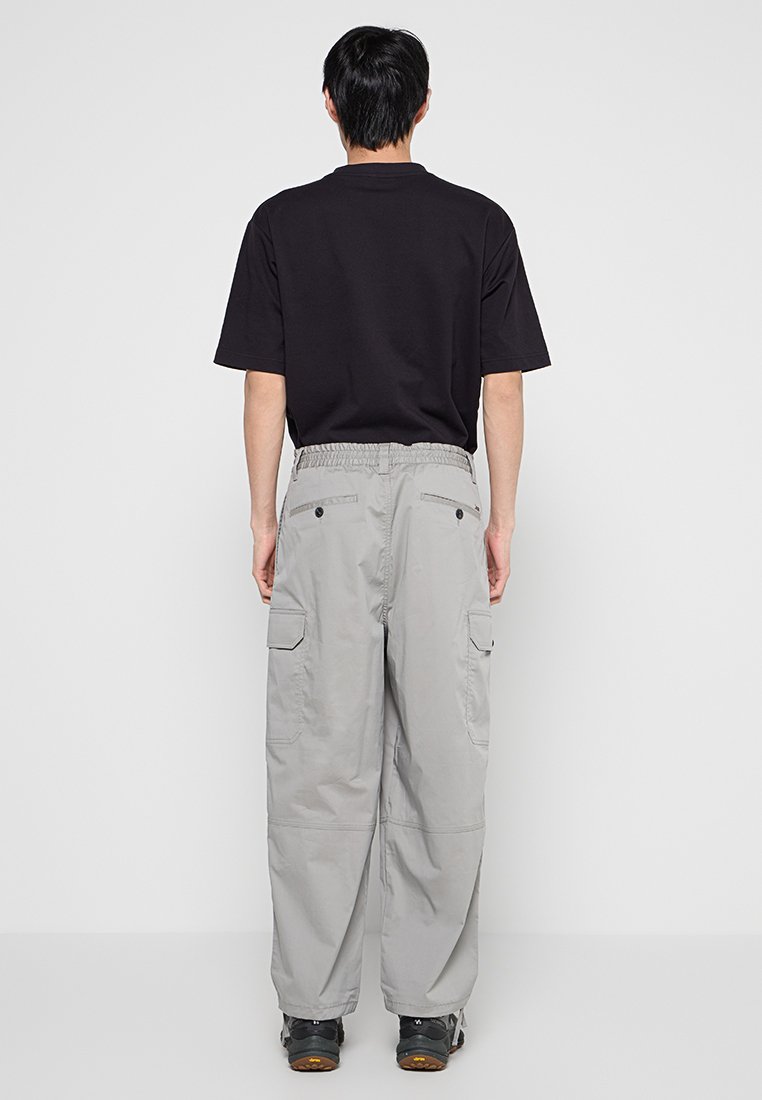 Hombre de pie de espaldas, vistiendo una camiseta negra de manga corta, pantalones cargo gris claro y zapatillas negras, sobre un fondo blanco liso.