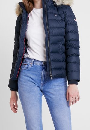 Femme portant une doudoune bleu marine avec capuche en fourrure, t-shirt blanc et jean bleu clair, debout devant un fond uni.