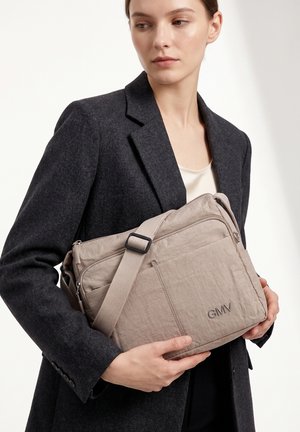 Giovane donna in blazer scuro che tiene una borsa a tracolla beige con tasca frontale con cerniera e logo GMV, guarda di lato su sfondo bianco.