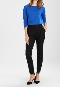 Blusa blu a maniche lunghe, vestibilità ampia, abbinata a pantaloni neri su misura. Scarpe col tacco nere, tessuto liscio, linee pulite, accenti minimi.