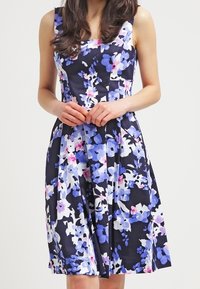 Vestido floral con fondo azul marino, adornado con flores azules, moradas y rosas. Presenta un cuerpo ajustado y una falda acampanada.