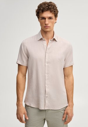 Młody mężczyzna z kręconymi włosami, ubrany w jasnobeżową koszulę z krótkim rękawem na guziki i jasnoszare spodnie, stojący na prostym tle.