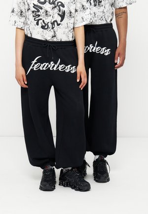 To personer iført sorte joggingbukser med "fearless" i hvid skrift og matchende sorte sneakers, står side om side på en hvid gulv.