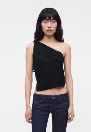 Ung kvinde med langt sort hår iført en asymmetrisk sort ærmeløs top og mørkeblå jeans, stående mod en ensfarvet baggrund.