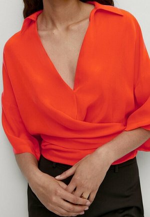 Femme portant un chemisier orange vif avec un col en V profond et un pantalon noir, les mains légèrement jointes devant sa taille.