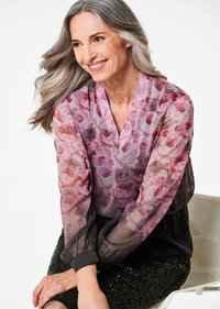 Durchsichtige Bluse mit pinkem Rosenmuster, V-Ausschnitt und schwarzen Bündchen. Getragen mit einem glitzernden schwarzen Rock. Glattes, leichtes Material.