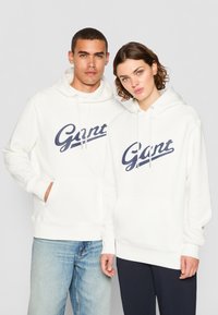Hvite hettegensere laget av myk stoff, med en stor marineblå "Gant" logo, kenguru-lommer og snorer på hettene.