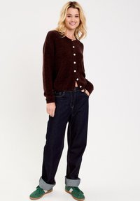 Cardigan di lana marrone con bottoni bianchi, abbinato a jeans blu scuro con risvolto e sneakers verdi. Tessuto morbido e vestibilità casual.