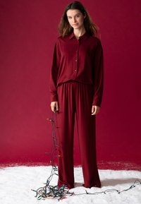 Bordeaux knoopshirt en bijpassende elastische broek, beide gemaakt van zacht materiaal. Model houdt een snoer met veelkleurige kerstverlichting vast.