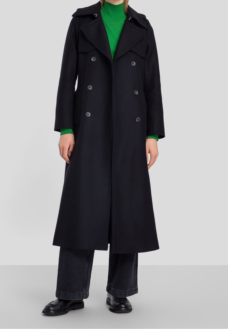 Cappotto nero doppiopetto con un design strutturato, grandi revers e spalline. Indossato sopra un maglione verde e pantaloni scuri.