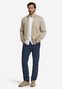 Beige cardigan över en vit skjorta, i kombination med mörkblå jeans och beige skor. Cardiganen har en slät textur och öppen framdelsdesign.