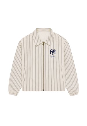 Ung mand iført en cremefarvet jakkesæt med striber, rødt bandana, mørkeblå kasket, lysegrå shorts, hvide sokker og sølvfarvede sneakers på en ensfarvet baggrund.