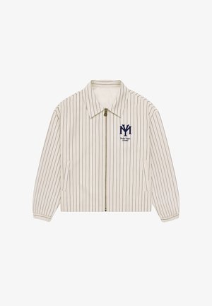 Ung mand iført en cremefarvet jakkesæt med striber, rødt bandana, mørkeblå kasket, lysegrå shorts, hvide sokker og sølvfarvede sneakers på en ensfarvet baggrund.