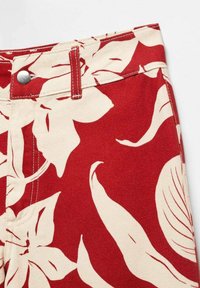 Pantalon taille haute rouge et crème avec grand imprimé floral, bouton argenté, passants pour ceinture et coutures visibles à la taille.