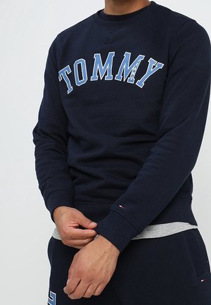 Mężczyzna poprawiający rękaw granatowej bluzy Tommy Jeans z białymi i jasnoniebieskimi napisami, w zestawie z granatowymi spodniami.
