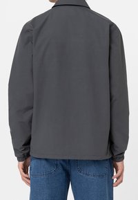 Grå lättjacka med kragdesign, elastiska ärmslut och en slät textur, bärs över blå denimbyxor. Bakifrån.