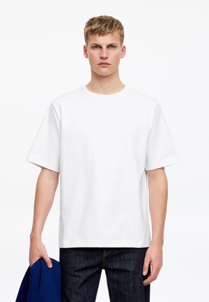 Jeune homme aux cheveux blonds courts portant un t-shirt blanc uni et un jean foncé, tenant un vêtement bleu plié dans sa main gauche.