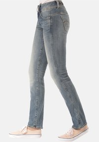 Kvinna iklädd urtvättade blå skinny jeans och beige canvas sneakers står på sidan mot en vit bakgrund.