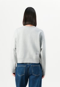 Persona con capelli neri lisci indossa una felpa grigia chiara e jeans blu a vita alta, di spalle su uno sfondo bianco.