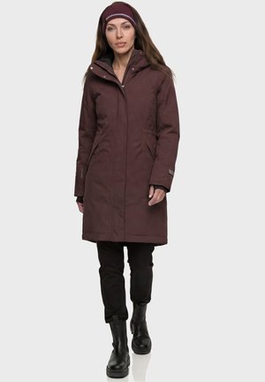 Frau trägt einen langen braunen Wintermantel mit Kapuze, schwarze Hose, schwarze Stiefel und ein gestricktes dunkelrotes Stirnband vor einfachem Hintergrund.