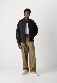 Man standing with hands in pockets wearing black bomber jacket, white ribbed top, wide khaki pants, and beige sneakers on plain background.