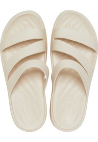 Beżowe klapki typu slip-on z trzema szerokimi, miękkimi pasekami, wytłaczanym wkładem oraz gładką fakturą. Na wkładzie widnieje wytłoczone logo marki "Crocs".