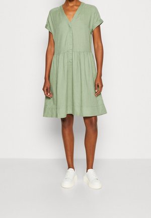 Personne portant une robe courte à manches courtes, de couleur vert clair, longueur genou, avec un décolleté en V et des baskets compensées blanches, se tenant devant un fond uni.