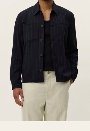 Homme portant une veste noire à rayures fines avec boutons sur une chemise noire et un pantalon beige, debout avec une main dans la poche.