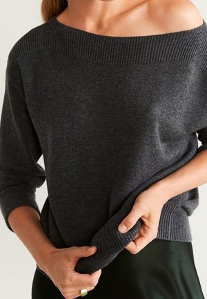 Femme portant un pull gris foncé décolleté aux épaules avec un col et des poignets côtelés, tenant l'ourlet du pull avec les deux mains.