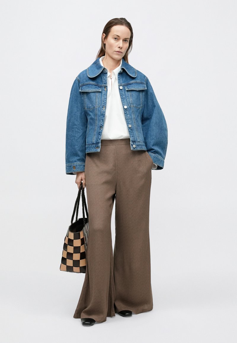 Denim jacket with a large collar, white blouse, and wide brown trousers. Holding a checked black and beige bag; black shoes visible.