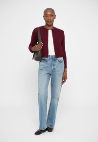 Bordeaux gebreide cardigan met een ronde hals, knoopsluiting en frontzak, gecombineerd met lichtblauwe flared jeans en zwarte schoenen.