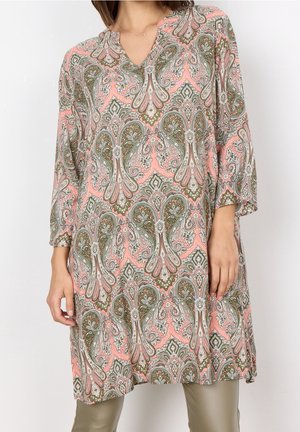 Kvinde iført langærmet tunika med pink, grønne og beige paisley-mønster, kombineret med lyse olivengrønne bukser, mod en ensfarvet hvid baggrund.