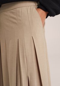 Gonna plissettata beige con cuciture visibili e tasca laterale, indossata con un top scuro a maniche lunghe, mano posata casualmente nella tasca.