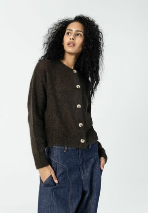 Mujer con cabello rizado, vestida con un cárdigan marrón oscuro abotonado y jeans azules, con las manos en los bolsillos, mirando ligeramente hacia la izquierda sobre un fondo blanco.