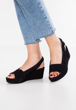 Voeten met zwarte suède sleehak sandalen met open tenen en enkelbandjes, gecombineerd met korte lichtblauwe jeans.