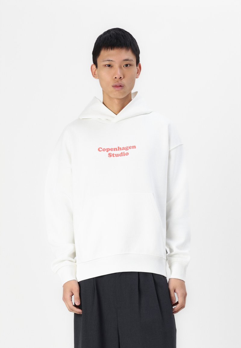 Witte hoodie met "Copenhagen Studio" in het roze op de borst, voorzien van een grote zak en een trekkoordcapuchon. Het materiaal lijkt zacht en glad te zijn.