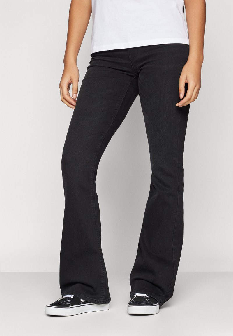 Sorte flared denim jeans med en glatt tekstur, tettsittende over livet og lårene, som flareer fra kneet til kanten. Brukt med sorte sneakers.