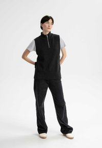 Schwarzes, geripptes, ärmelloses Oberteil mit einem hohen Kragen und Reißverschluss, kombiniert mit einem grauen T-Shirt mit kurzen Ärmeln und schwarzen Cordhosen. Helle Schuhe.