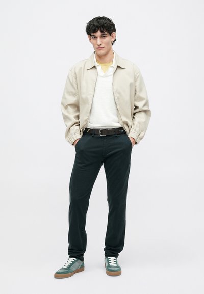 Šviesiai rudos bomberio striukė užsidėta ant baltos megztinio, žalios kelnės ir žalios sportbačiai su rudais akcentais. Modelis stovi prieš paprastą baltą fono.