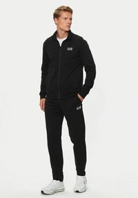 Giacca sportiva nera con zip e pantaloni abbinati, entrambi realizzati in materiale morbido. Presenta il logo sul petto e sulla coscia. Sneakers bianche completano il look.