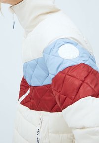 Prošívaná péřová bunda v krémové, světle modré a vínové barvě s vysokým límcem, zipem a logem na levém rukávu. Hladká a teplá textura.