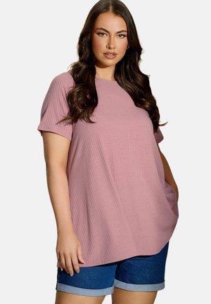 Kobieta z długimi ciemnymi włosami, ubrana w różowy, prążkowany top z krótkim rękawem i granatowe, podwinięte szorty jeansowe, stoi z jedną ręką w kieszeni.