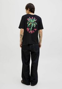 Sort t-shirt med et stort, flerfarvet palmetræ-grafik og lyserød tekst "PLAZA BLANCA" på ryggen. Bæret med mørkeblå denim bukser.
